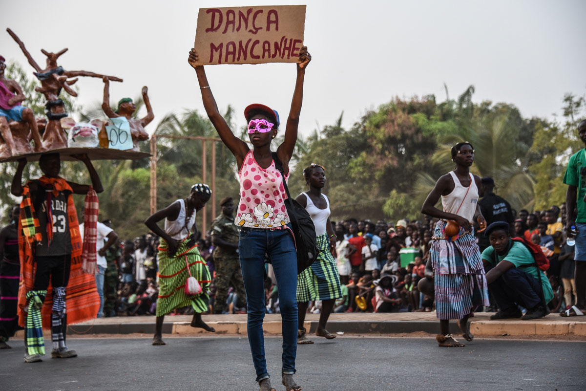 A woman from Bissau holds up a sign announcing her troupe's performance of a 'Mancanhe dance', as they parade down the streets of the capital. [Ricci Shryock/Al Jazeera]